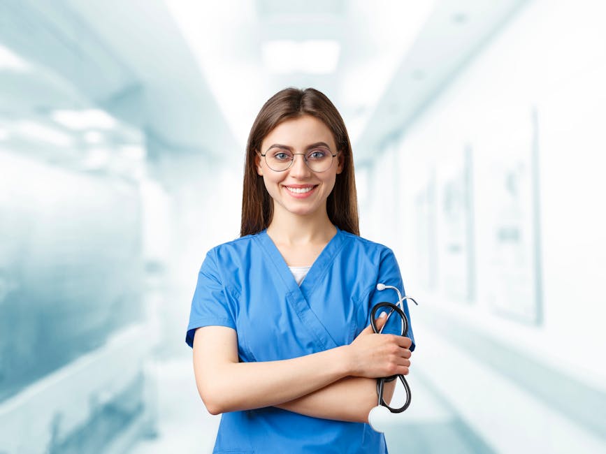 Young nurse in blue scrubs smiling and holding a stethoscope in a hospital corridor