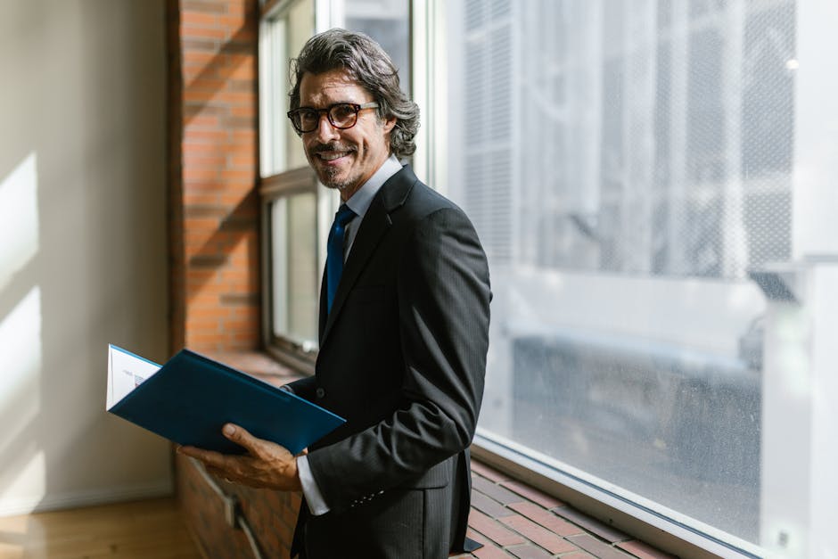 Smiling businessman in a black suit holding a folder by a window indoors